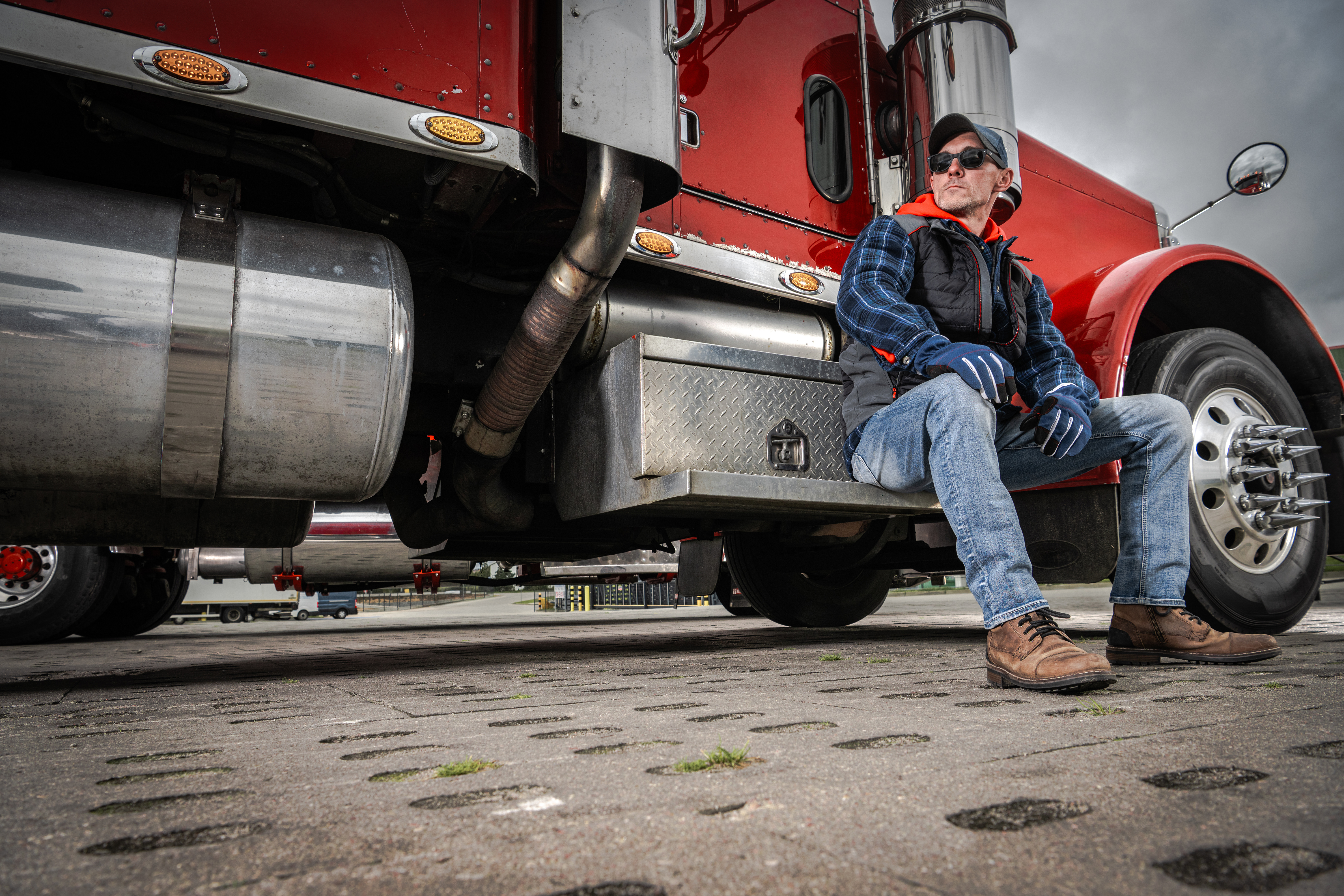 Truck driver in blue shirt and cap sitting on red semi-truck cab step during daytime