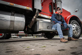 Truck driver in blue shirt and cap sitting on red semi-truck cab step during daytime