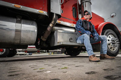 Truck driver in blue shirt and cap sitting on red semi-truck cab step during daytime