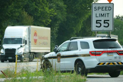 Police SUV parked on roadside near speed limit 55 sign with delivery truck and trees in background
