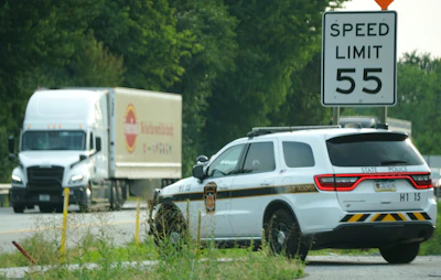 Police SUV parked on roadside near speed limit 55 sign with delivery truck and trees in background
