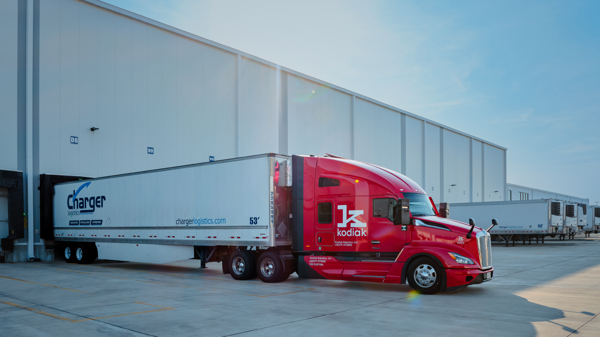 Red Kodiak autonomous truck with Charger Logistics trailer parked at industrial warehouse facility under clear sky