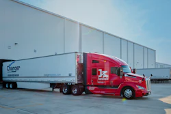 Red Kodiak autonomous truck with Charger Logistics trailer parked at industrial warehouse facility under clear sky