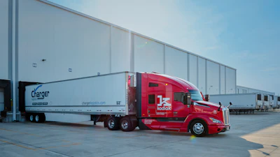 Red Kodiak autonomous truck with Charger Logistics trailer parked at industrial warehouse facility under clear sky