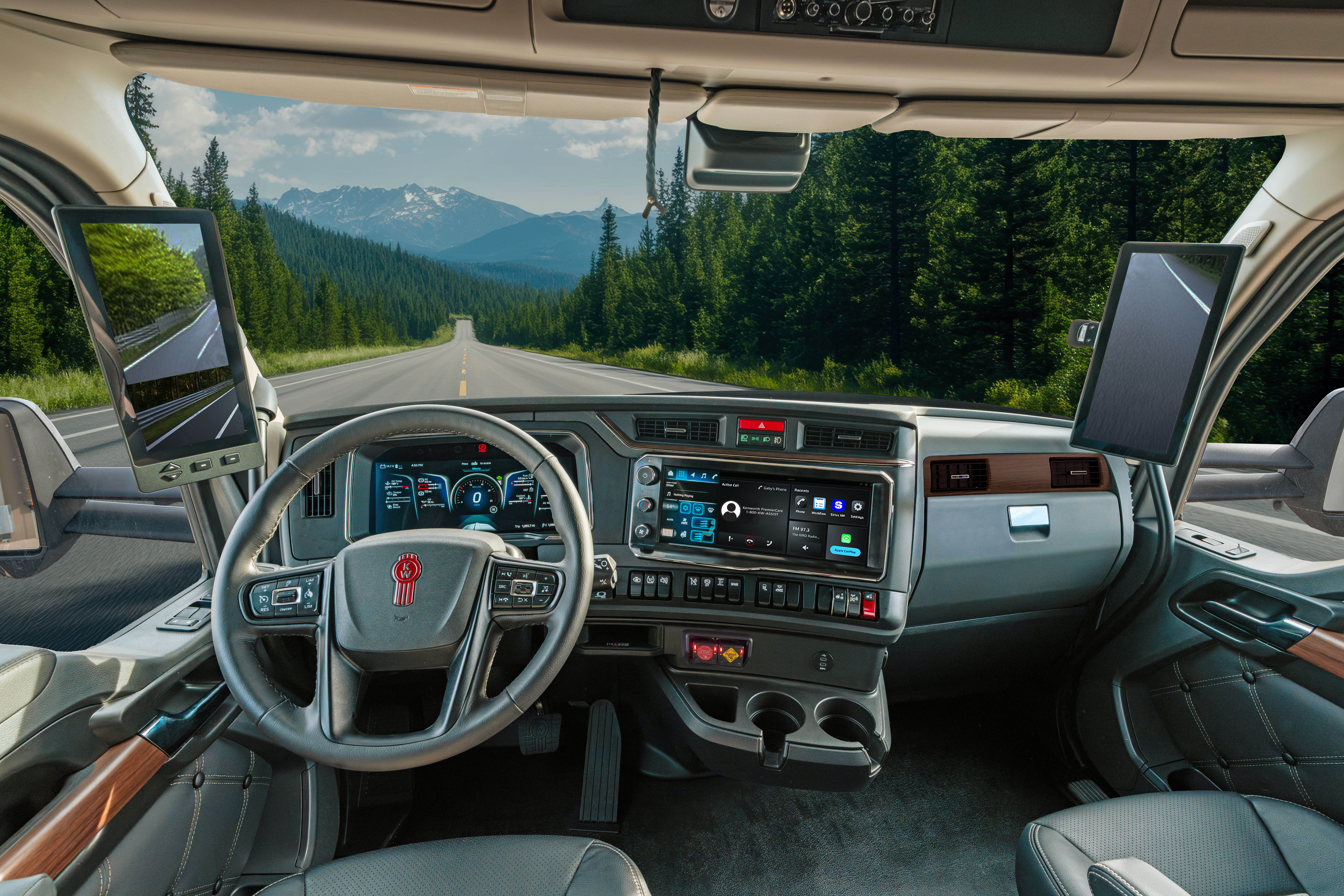 Interior view of a truck cab with steering wheel and dashboard, looking out at a straight mountain highway lined with evergreen forests.