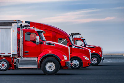 Three red commercial semi-trucks lined up on a paved surface against a cloudy sky