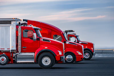 Three red commercial semi-trucks lined up on a paved surface against a cloudy sky