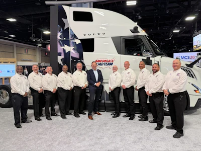 Professional truck drivers in white shirts pose in front of a Volvo semi-truck branded with America's Road Team logo at an indoor trade show.