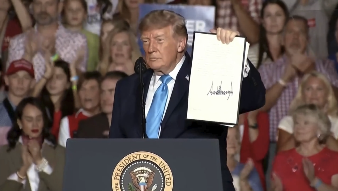 Person at podium holding document during political rally with crowd in background