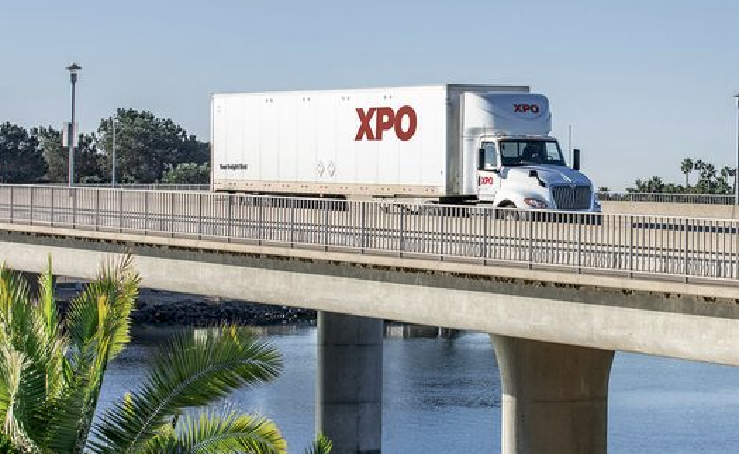 XPO Logistics branded semi-truck traveling on elevated highway bridge over water with palm trees visible below