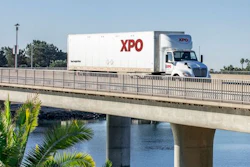 XPO Logistics branded semi-truck traveling on elevated highway bridge over water with palm trees visible below