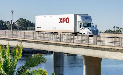XPO Logistics branded semi-truck traveling on elevated highway bridge over water with palm trees visible below