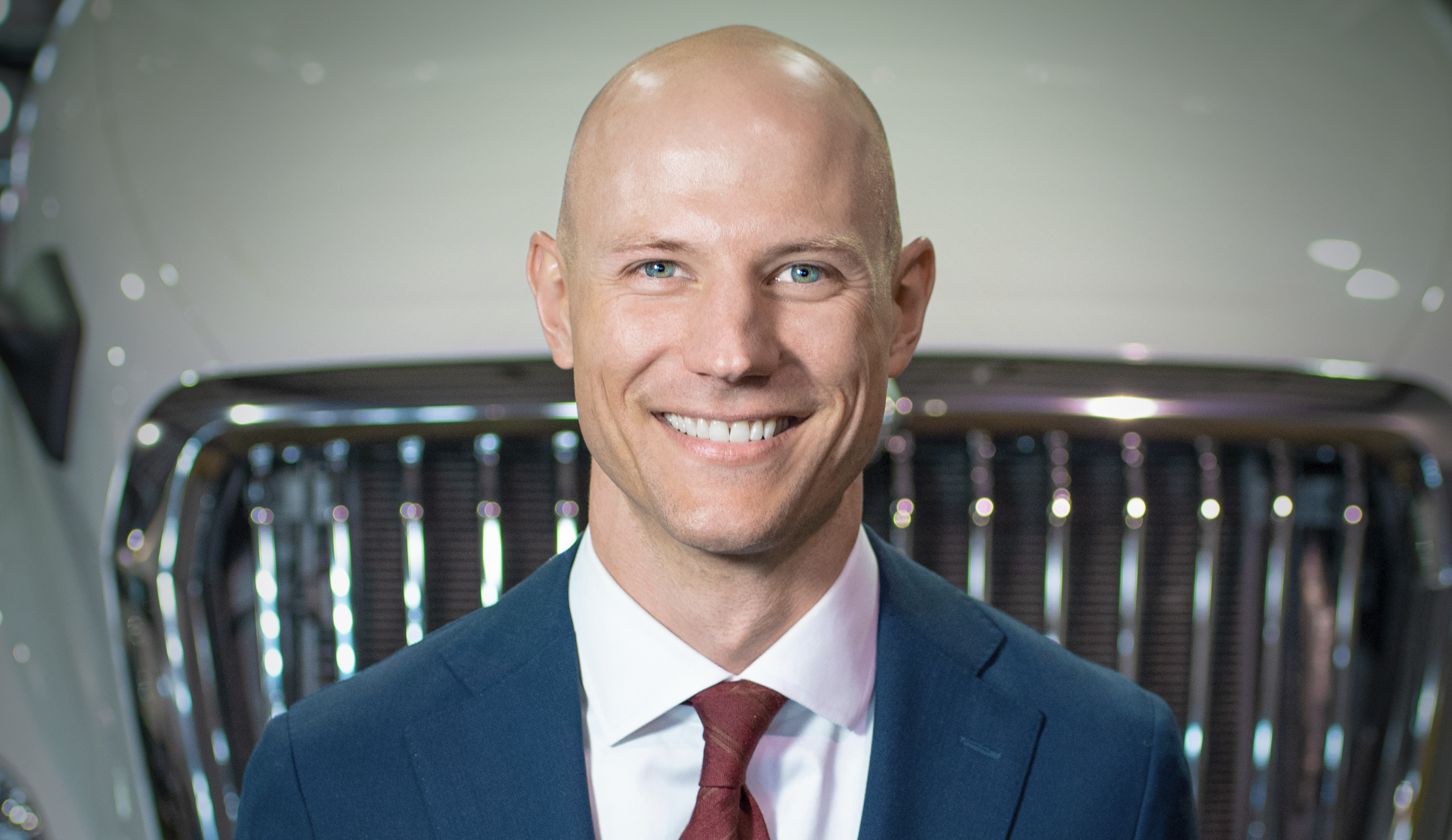 Professional headshot of a smiling man in navy blazer and burgundy tie against automotive showroom background