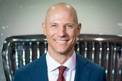 Professional headshot of a smiling man in navy blazer and burgundy tie against automotive showroom background