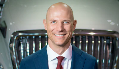 Professional headshot of a smiling man in navy blazer and burgundy tie against automotive showroom background