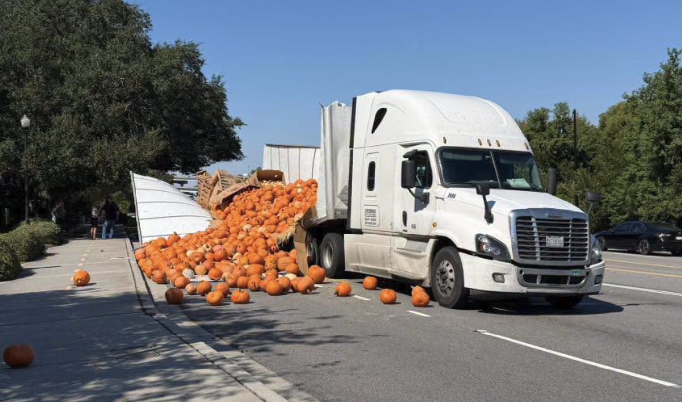 White semi-truck with overturned trailer spilling orange pumpkins across parking lot