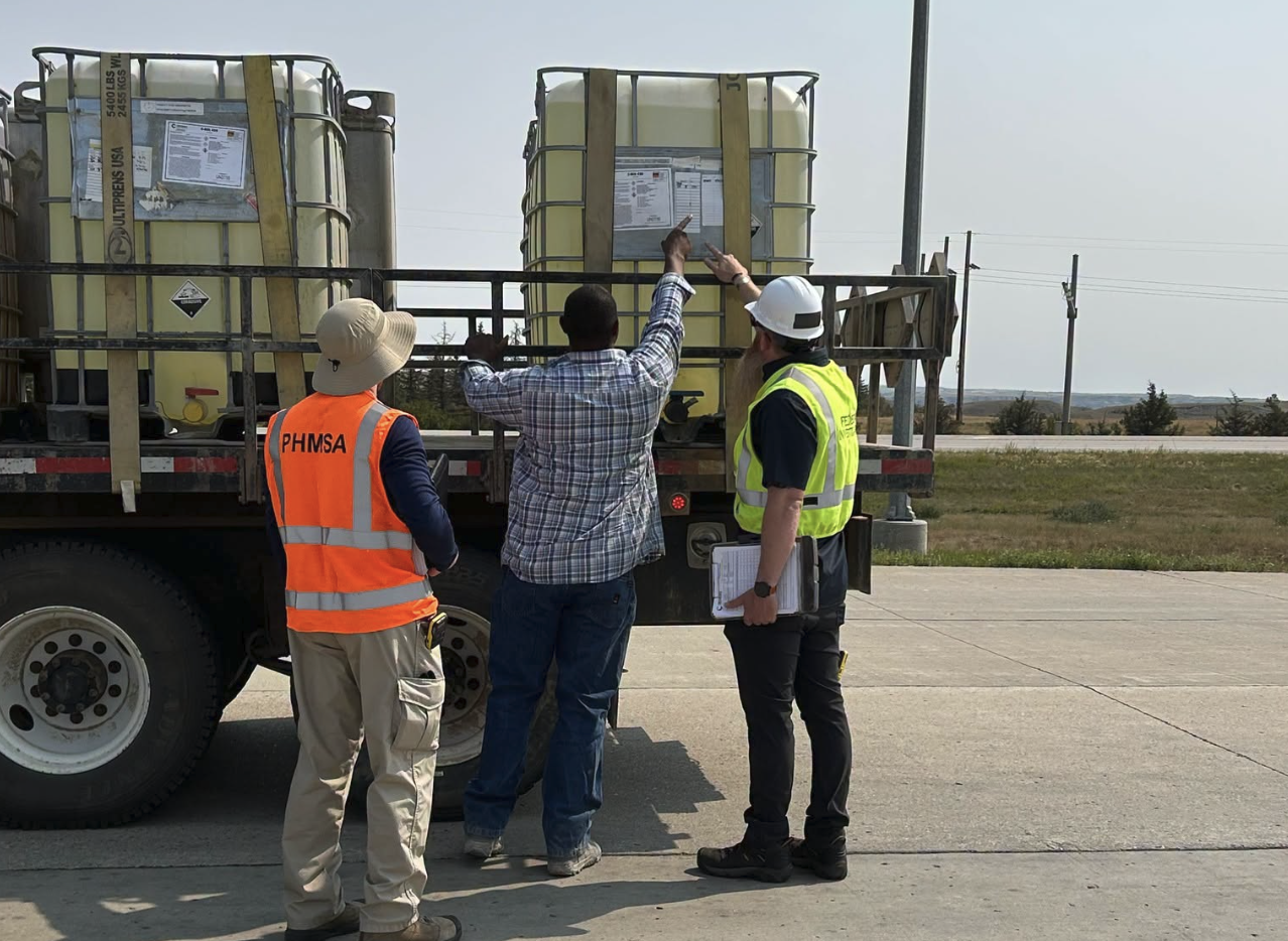 Three workers in safety vests inspect large industrial storage containers on a truck bed at an outdoor facility.