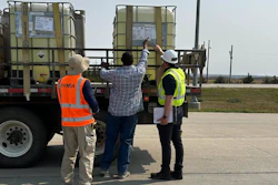Three workers in safety vests inspect large industrial storage containers on a truck bed at an outdoor facility.