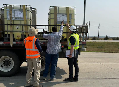 Three workers in safety vests inspect large industrial storage containers on a truck bed at an outdoor facility.