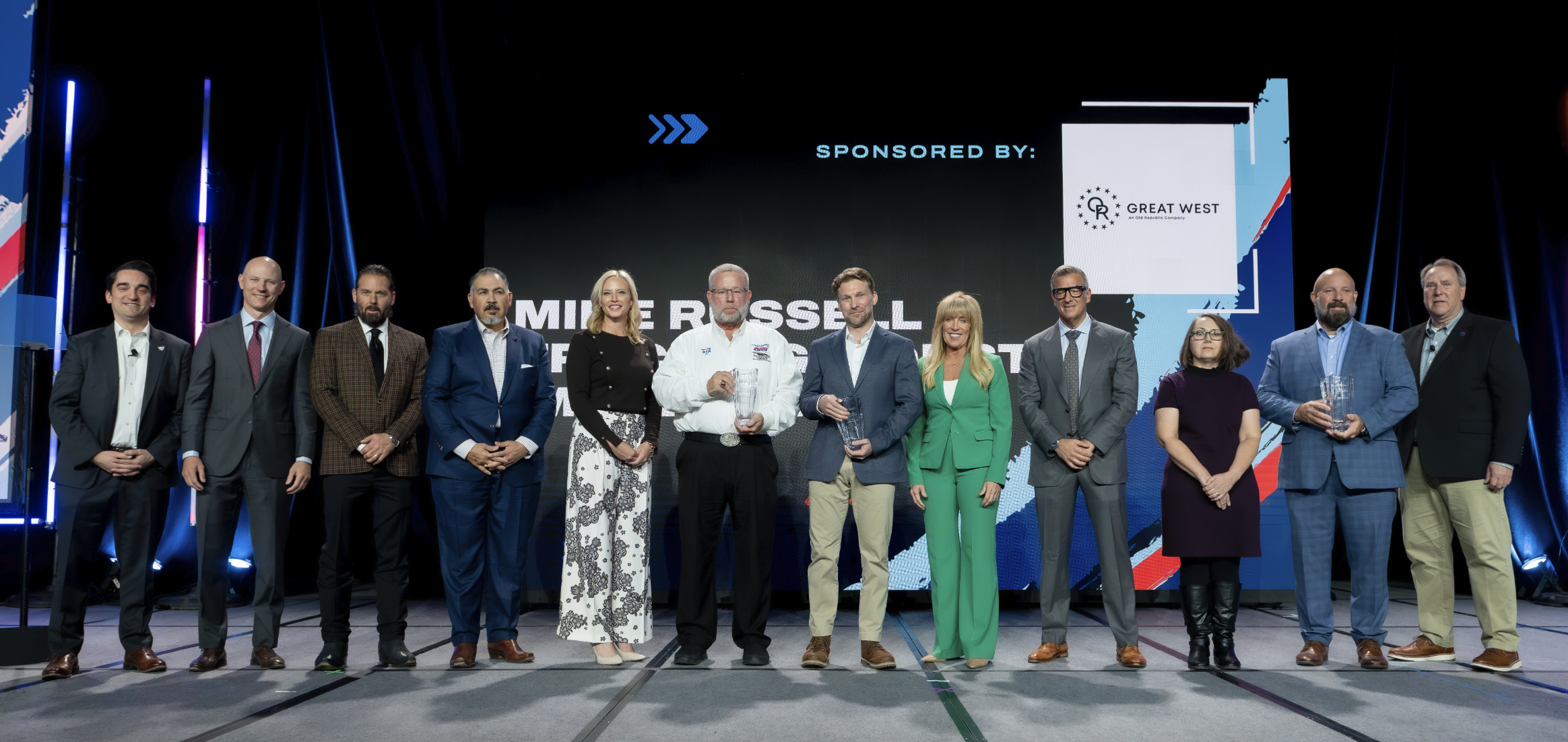Group of business professionals standing on stage at formal event with Great West sponsorship banner