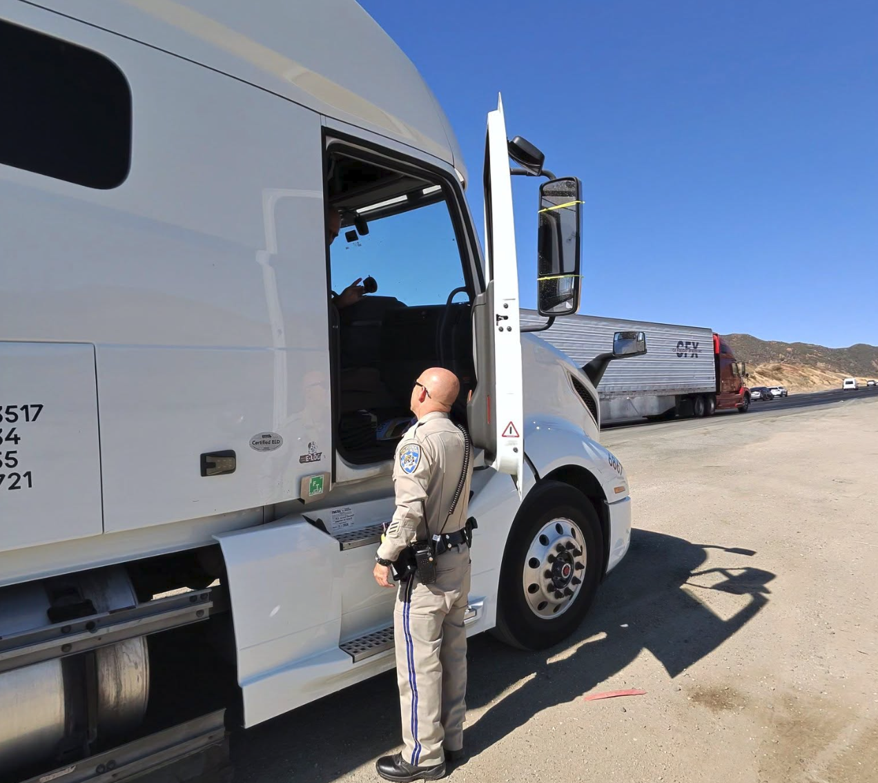 Law enforcement officer inspecting commercial truck cab at roadside checkpoint under clear sky