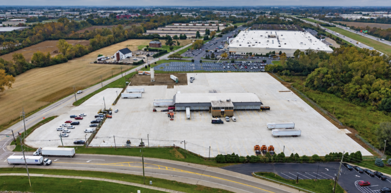 Aerial view of industrial facility with white-roofed buildings, parking lots, and surrounding farmland near highway