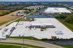 Aerial view of industrial facility with white-roofed buildings, parking lots, and surrounding farmland near highway