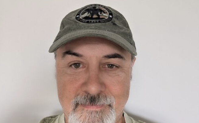 Middle-aged man with gray beard wearing olive green cap with emblem, neutral expression against white background