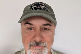 Middle-aged man with gray beard wearing olive green cap with emblem, neutral expression against white background