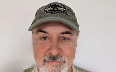 Middle-aged man with gray beard wearing olive green cap with emblem, neutral expression against white background