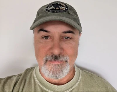 Middle-aged man with gray beard wearing sage green cap and t-shirt against neutral background