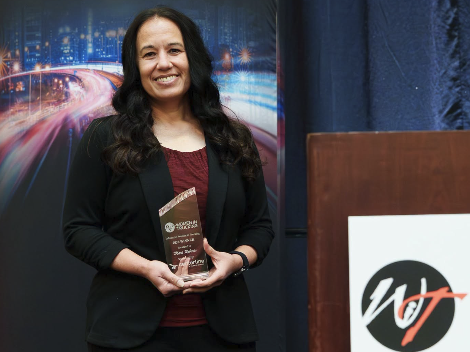 Woman in black blazer holding award at WT organization event with cityscape backdrop