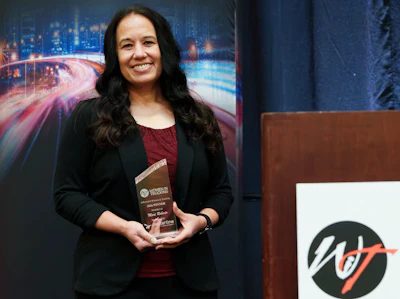 Woman in black blazer holding award at WT organization event with cityscape backdrop