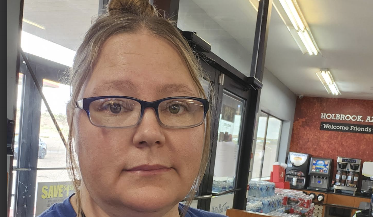 Woman wearing blue-framed glasses in a casual indoor setting with storefront signage visible in the background