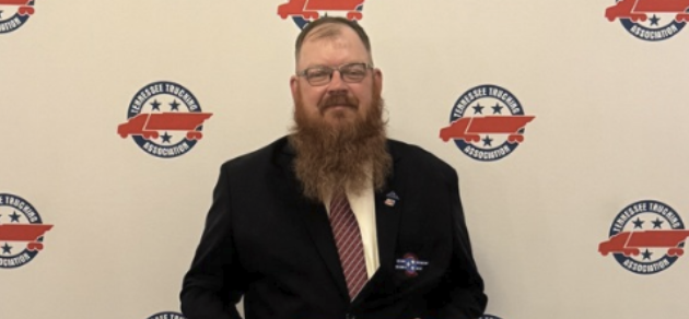 Professional man with beard and glasses wearing dark blazer at formal event with branded backdrop