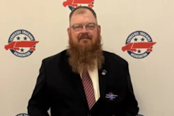 Professional man with beard and glasses wearing dark blazer at formal event with branded backdrop