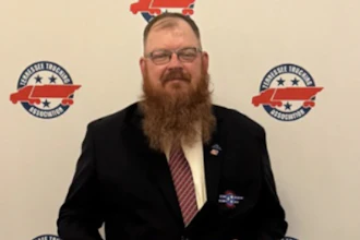 Professional man with beard and glasses wearing dark blazer at formal event with branded backdrop