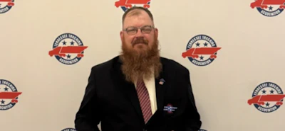 Professional man with beard and glasses wearing dark blazer at formal event with branded backdrop