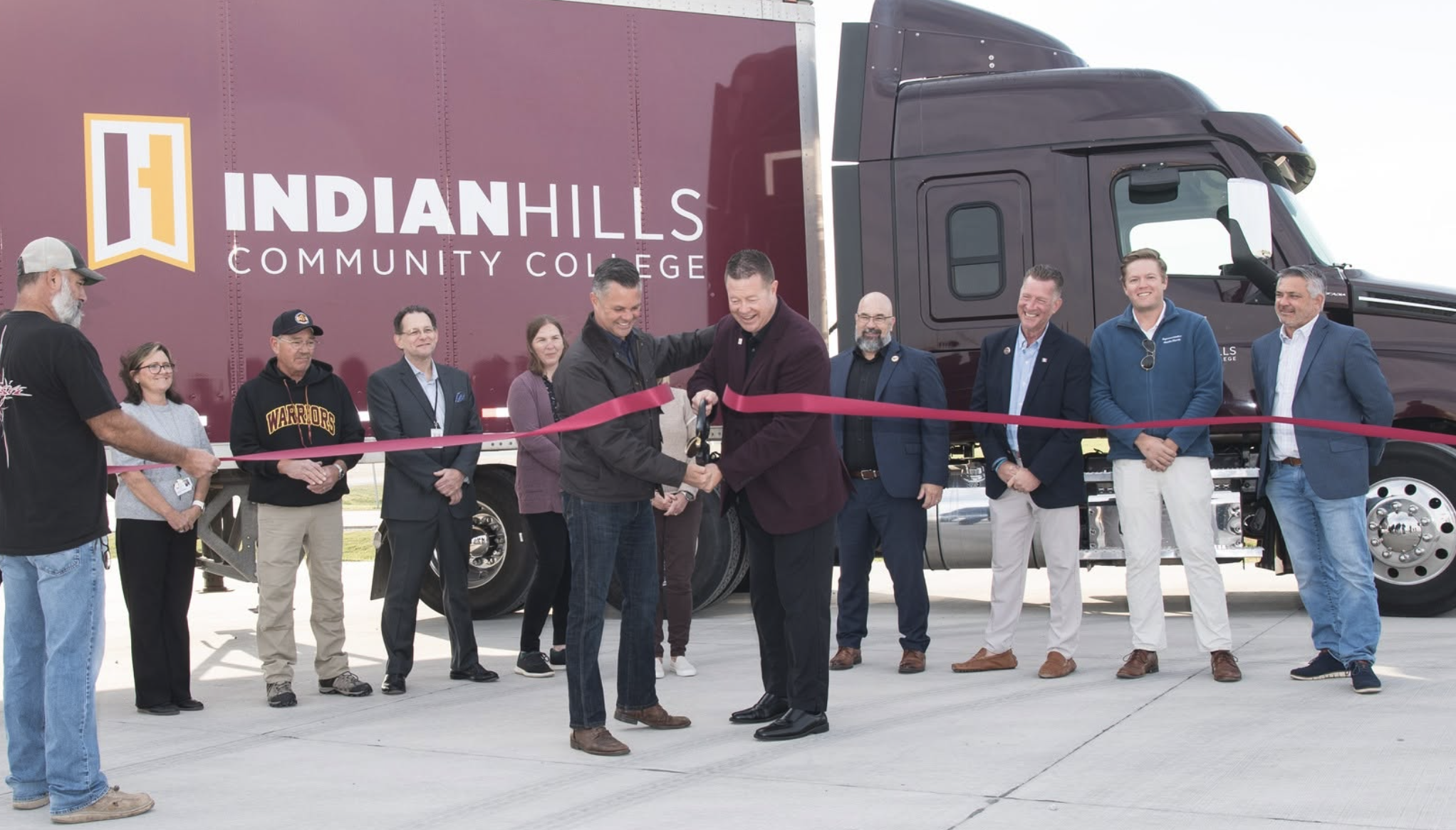 Group of officials and community members participate in ribbon-cutting ceremony at Indian Hills Community College event with branded vehicle