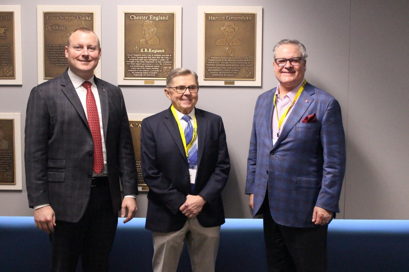 Three professional men in business attire standing together in front of commemorative bronze plaques on a wall.