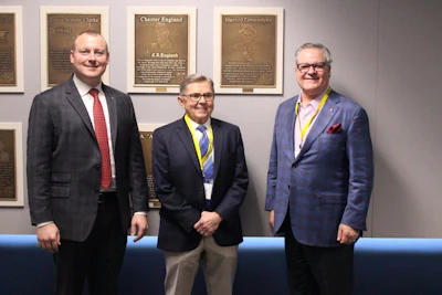 Three professional men in business attire standing together in front of commemorative bronze plaques on a wall.